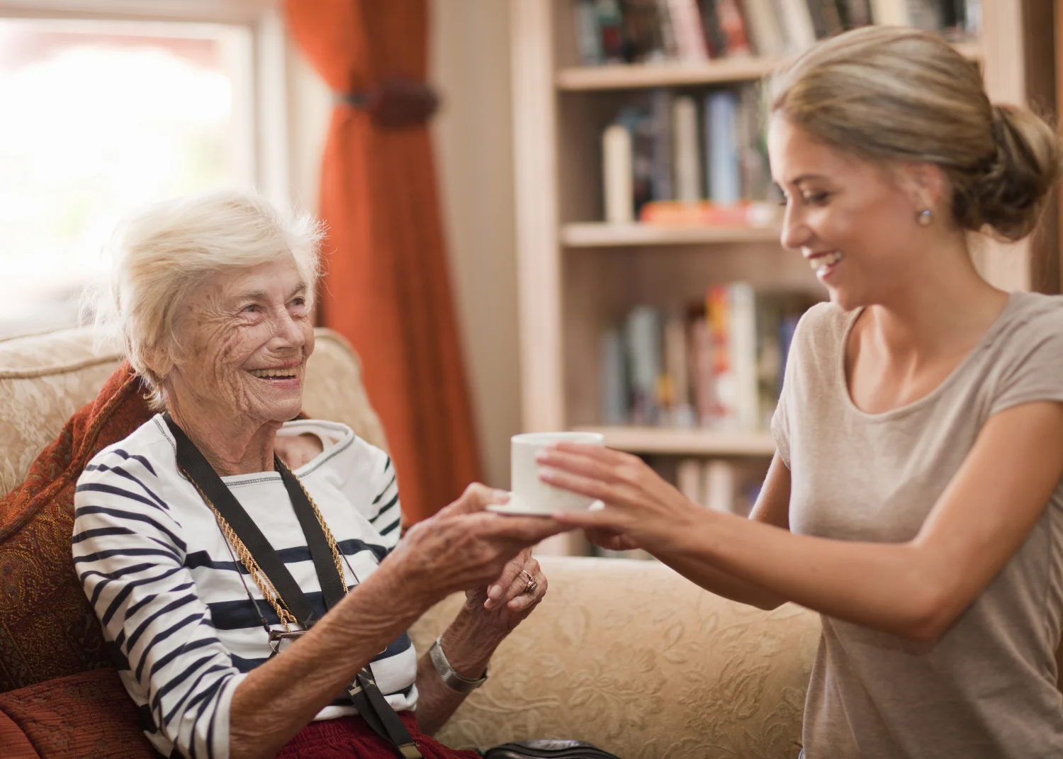 Care assistant handing coffee to smiling senior woman.